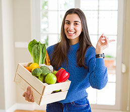 Beautiful young woman holding wooden box full of healthy groceries very happy pointing with hand and finger to the side